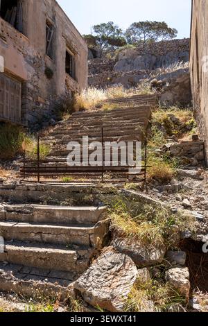 Weathered concrete staircase with metal railing stands between ruined stone buildings with green trees on hill under clear sky on Spinalonga island Cr Stock Photo