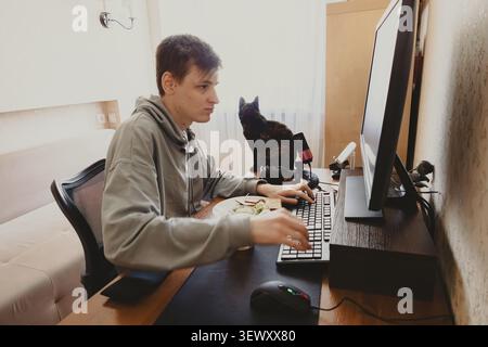 Young person working on computer at home desk with cat companion Stock Photo