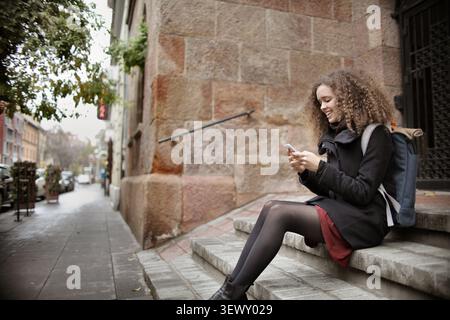 Happy woman with curly hair using phone on city steps Stock Photo
