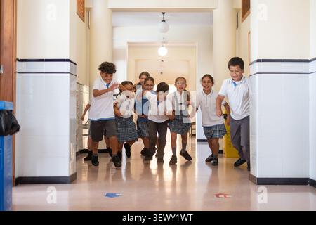 Group of cheerful elementary school students running together down a bright school corridor during a break. Stock Photo