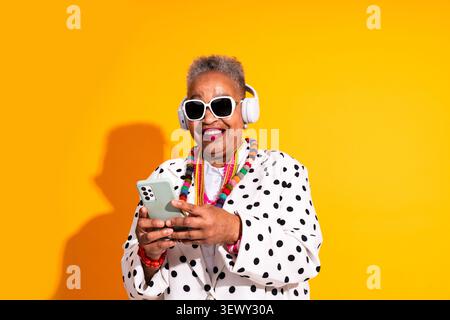 Black senior woman wearing modern clothing portrait on background - happy senior woman wearing white headphones, sunglasses, and polka dot shirt, hold Stock Photo