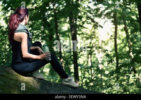 Woman with red hair sitting in a dark green forest, relaxing, holding a water bottle Stock Photo