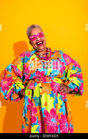 Black senior woman wearing modern clothing portrait on background - joyful senior black woman wearing bright floral dress and pink glasses, standing c Stock Photo