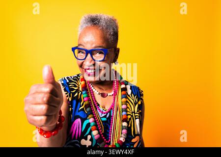 Black senior woman wearing modern clothing portrait on background - happy senior woman showing thumbs up, expressing positivity, satisfaction, and agr Stock Photo