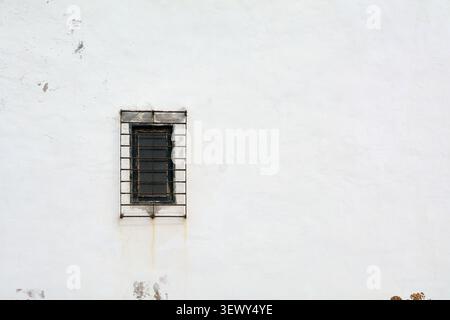 A small old window with rusty metal bars set into a large, weathered white stucco facade of a traditional house in Agaete. Stock Photo