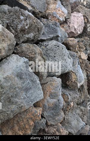 A close-up vertical view of a textured dry stone wall constructed from dark grey, brown, and reddish porous volcanic lava rocks in Agaete. Stock Photo