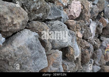 A close-up perspective view of a rugged, textured stone wall constructed from dark grey and brown volcanic lava rocks in Agaete. Stock Photo