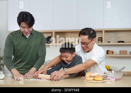 Happy Asian grandfather and father helping boy roll dough in kitchen Stock Photo