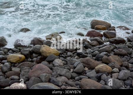 Foamy sea water ripples and splashes against a collection of large, rounded coastal rocks and boulders on a shore in Agaete, Spain. Stock Photo
