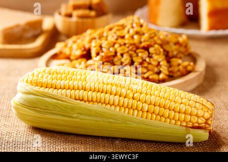 Boiled corn on the cob on a table featuring traditional Brazilian foods, with homemade pe de moleque in the background. Traditional cuisine. Stock Photo