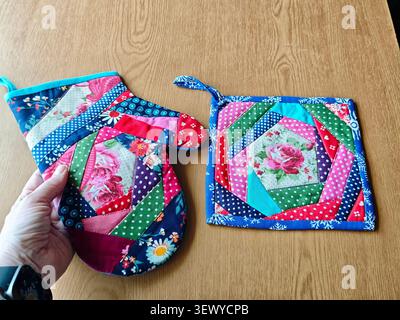 A person holds a quilted oven mitt while a pot holder rests on the table. Both have bright patterns and floral designs. Stock Photo