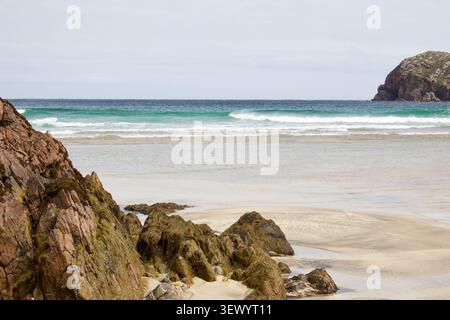 Weathered rust-coloured rocks frame a calm turquoise shoreline, with a distant headland visible under soft overcast sky at Cliff Beach, Isle of Lewis Stock Photo