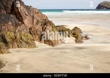 Weathered, lichen-covered rocks emerge from wind-rippled sand in foreground, with turquoise waves breaking beneath a distant headland at Cliff Beach Stock Photo