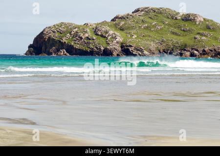 A turquoise wave curls beneath a twin-peaked rocky headland, with wide sand flats stretching across the foreground at Cliff Beach, Isle of Lewis, Oute Stock Photo