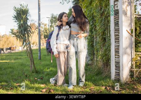 Two girls are standing together in a park. One girl leans against the other while they smile and talk. Trees and grass are around them in the backgrou Stock Photo