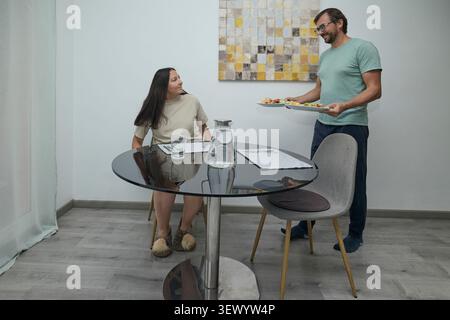 Couple having lunch at home. Man and woman sitting at the dining table, serving and eating healthy homemade food. Concept of domestic life, togetherne Stock Photo