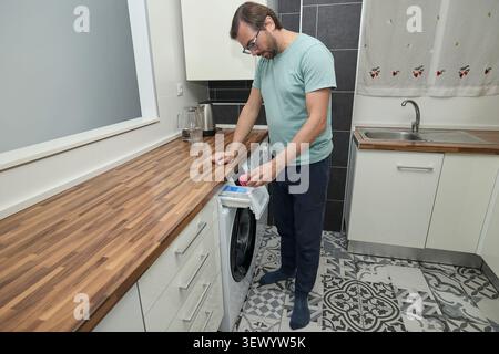 Man doing laundry at home in modern kitchen. Person loading washing machine and pouring detergent. Concept of household chores, domestic life, and hom Stock Photo