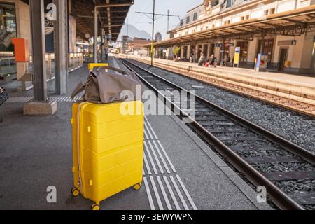 Bright yellow suitcase standing on a railway platform beside train tracks. Travel background with copy space for tourism, transportation advertising Stock Photo