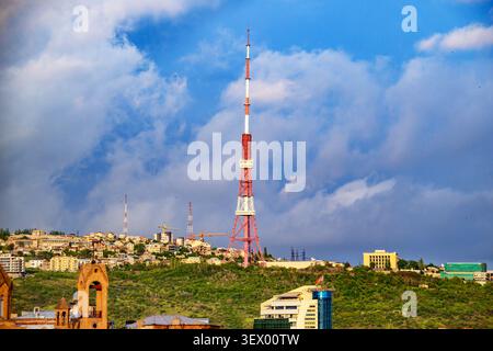 Tall red-and-white broadcast tower rises above green hillside town. Blue sky with white clouds dominates background. Buildings scattered across hillto Stock Photo