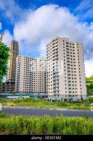 Tall residential buildings stand against bright blue sky with fluffy clouds. Green grass and trees grow near asphalt road in foreground. One building Stock Photo