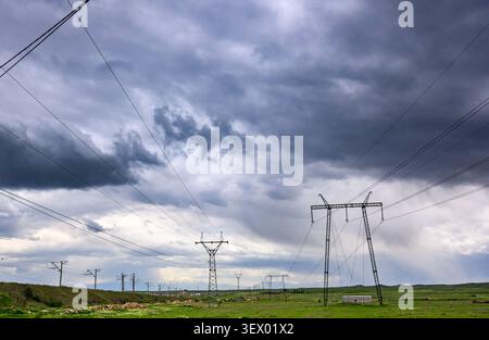 Dark storm clouds loom over green field. Power lines stretch across landscape. Metal pylons stand tall among grassy terrain Stock Photo