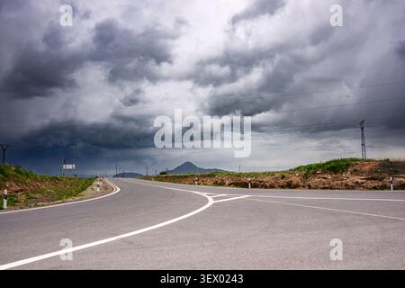 Winding road curves under dark storm clouds. Grassy shoulders flank asphalt path. Utility poles line horizon. Distant hill peaks emerge through mist Stock Photo