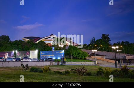 Modern stadium at dusk, lit by streetlights. Grass and shrubs in foreground. Trees line pathway to venue. Sky deep blue fading to dark Stock Photo