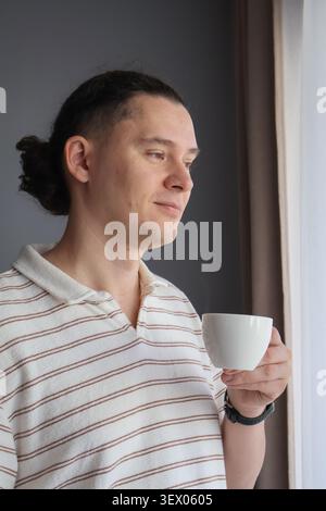 Relaxed middle aged man holding cup of coffee while looking through window at home. Quiet morning routine, relaxation, peaceful moment and comfortable Stock Photo