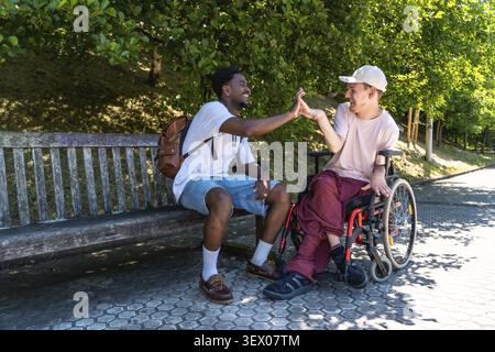 Two young diverse friends, one sitting on a bench and the other in a wheelchair, are sharing a happy moment in a park, reaching to give each other a h Stock Photo