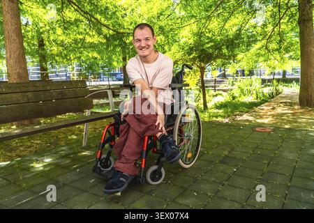 Young man with a disability confidently smiling and looking at the camera, sitting in a modern wheelchair outdoors in a vibrant green park on a bright Stock Photo