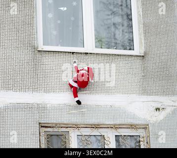 Santa Claus is hanging from a window sill. The scene is set in a building with a white facade Stock Photo