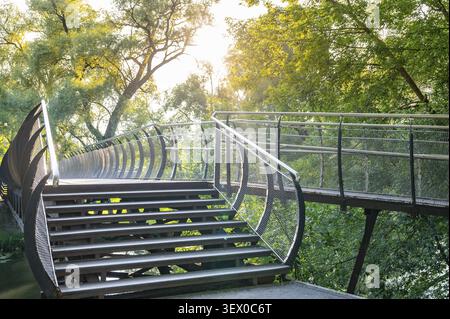 The curved footbridge with metal railings and steps is located in a lush green park where sunlight falls through the trees. The design blends seamless Stock Photo
