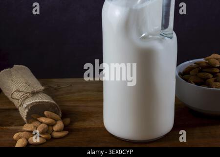 Almond milk in a clear glass bottle with whole almonds in a bowl and spilled from a burlap bag on a rustic wooden table Stock Photo