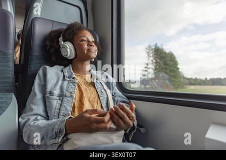 An African American woman sits in a train. She listens to music and looks out the window during her journey. Stock Photo