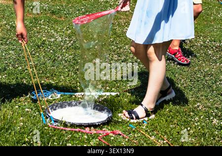 Children are engaged in blowing soap bubbles in a park, with one child holding a bubble wand and the other standing close by on the grass Stock Photo