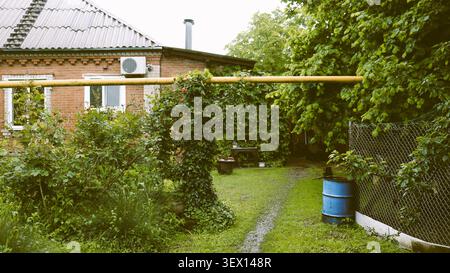 A village courtyard with a brick house. A yellow gas pipe, entwined with vines, stretches across a green area with bushes and a path Stock Photo