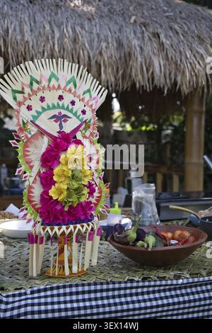Colourful Balinese offerings and fresh tropical fruits in a clay bowl in the background, a chef prepares food outdoors in front of a bamboo fence in B Stock Photo