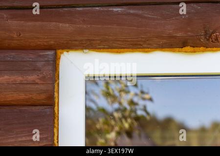 yellow polyurethane foam seal a white window frame corner on a brown timber log house exterior wall with reflection in glass. Stock Photo