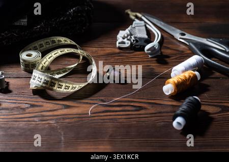 Scattered sewing threads on a wooden table with tailor scissors, measuring tape, thimble and clothing details. Stock Photo