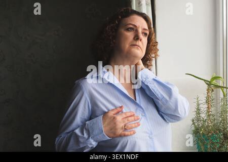 Pensive middle-aged woman holding her chest and neck by a window with houseplants, thoughtful and worried Stock Photo