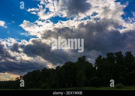 Dynamic sky with brewing storm clouds, vibrant blue patches, and sun rays breaking through. Dark, lush forest line below, adding depth to the dramatic Stock Photo