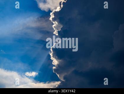 28 June 2026, Brandenburg, Sieversdorf: A massive, dark-blue thundercloud looms imposingly across the right half of the image, darkening the scene. The frayed edge of the dense wall of clouds is intensely backlit by the hidden sun, creating a dazzlingly bright silver streak. On the left, the view opens up to a deep-blue summer sky streaked with delicate veil-like clouds. Here and there, fine, fan-shaped rays of light break through the dark mass, tracing subtle paths across the atmosphere. This meteorological close-up captures the extreme contrast between bright sunshine and an approaching stor Stock Photo