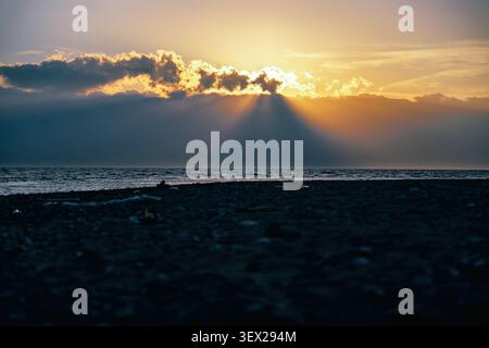 Dramatic sunset over the sea with sun rays breaking through clouds on a dark beach Stock Photo