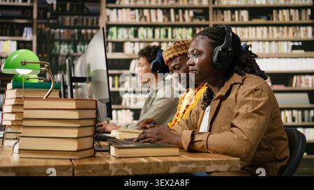 Smiling black student wearing headphones and typing at desktop monitor with books and classmates nearby in warm library studying space for coursework. Digital learning. Stock Photo