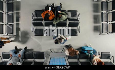 People on airport chairs talking next to passengers catching plane, hectic passenger movement concept. Travelers with trolleys chatting, waiting to be called to boarding gate, high angle view Stock Photo