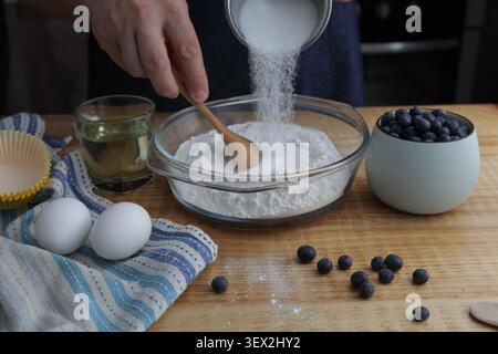 Person adding granulated sugar to flour while mixing ingredients for homemade blueberry muffins. Fresh blueberries, eggs and baking ingredients arrang Stock Photo