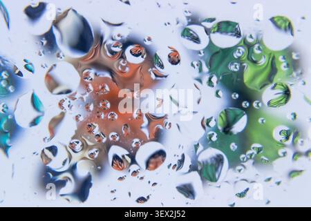 Raindrops cluster on a transparent surface. Reflections shimmer with warm, cool, and muted tones. Bubbles distort shapes and colors behind the glass. Stock Photo