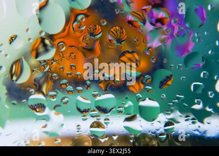 Raindrops distort vibrant, abstract color patterns on a glass surface with flowers. Tiny droplets form a shimmering, kaleidoscopic reflection. Colors Stock Photo