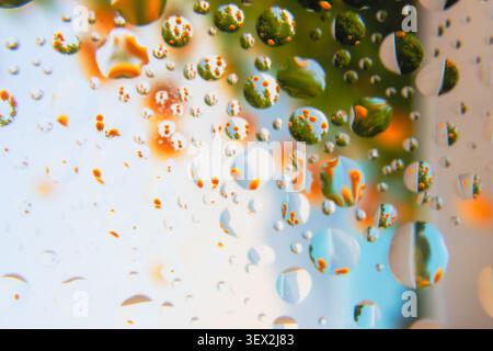 Raindrops cling to a glass surface. Reflections blur green and orange hues. Background hints at foliage, sky, and flowers. Dewy surface captures scatt Stock Photo