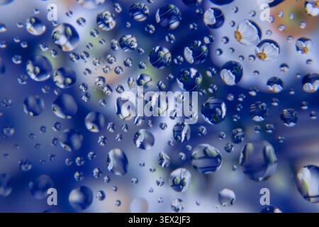 Blue-toned window with droplets reflecting blurred flower-like surroundings. Water beads cluster on glass surface. Background hints at soft, out-of-fo Stock Photo